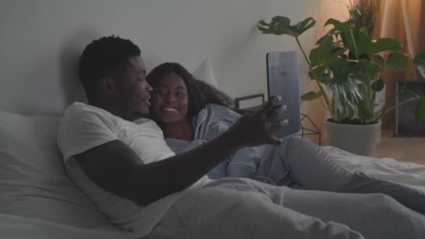 Цифровой телевизор. Счастливая африканская американская пара в любви отдыхает в постели и смотрит видео на цифровой планшет, отслеживая выстрел — стоковое видео