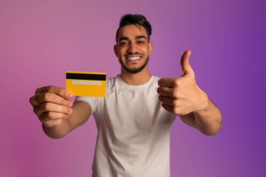Mutlu genç Arap adam elinde kredi kartı tutuyor ve neon ışıkta baş parmağını kaldırıyor, seçici odaklanıyor.