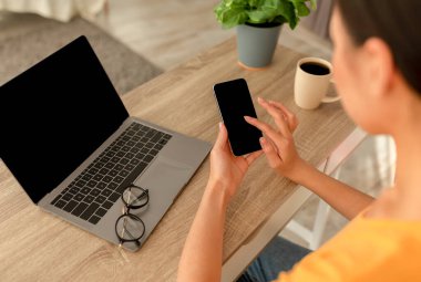 Tanımlanamayan genç bir kadın elinde boş ekranlı akıllı telefon, masada dizüstü bilgisayarlı, model şablonlu.