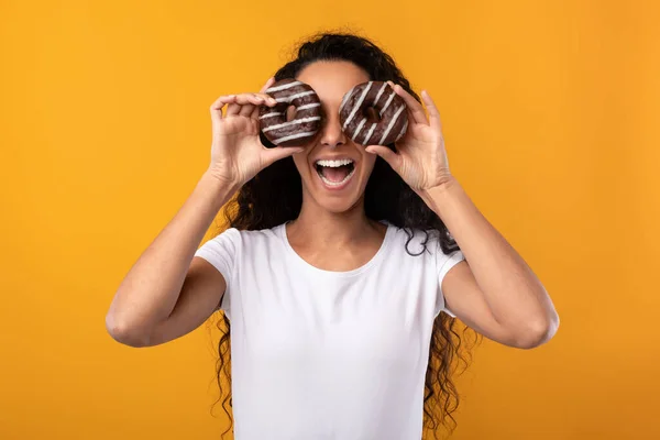 Feliz señora latina cubriendo los ojos con donuts en el estudio — Foto de Stock