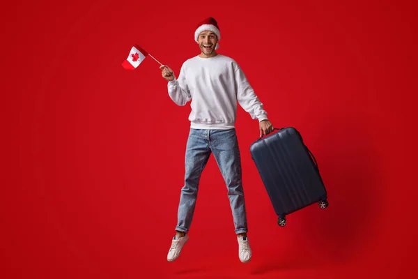 Чоловік, який носить Санта - Капелюх, стрибаючи в повітрі з канадським прапором і валізою — стокове фото
