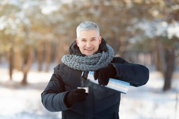 Hombre mayor sonriente en ropa de invierno caliente vertiendo té del termo en el parque nevado —  Fotos de Stock