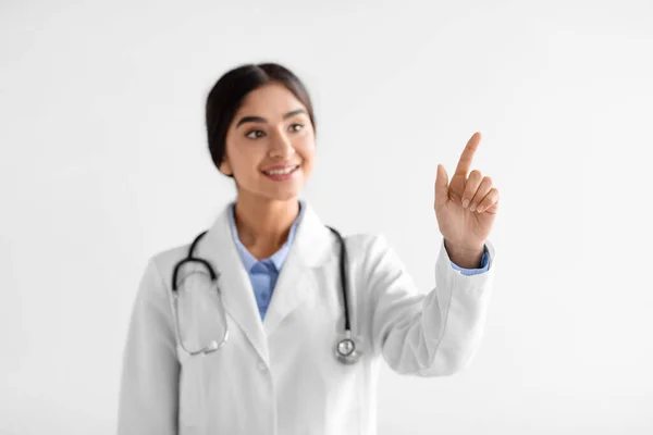 Sorrindo muito jovem médico mulher hindu em casaco branco com estetoscópio aponta dedo para espaço em branco — Fotografia de Stock