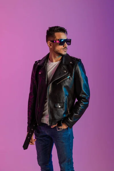 Retrato de joven árabe guapo en gafas de sol con jeans y chaqueta de cuero negro, posando en luz de neón — Foto de Stock