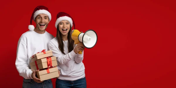 Tatil tanıtımı. Neşeli çift Noel Baba Şapka Giyiyor Megafon ve Hediye Kutuları Tutuyor — Stok fotoğraf