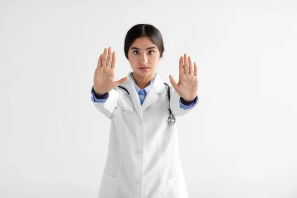 Serious joven india médico en abrigo con estetoscopio hace señal de stop con las manos sobre fondo blanco —  Fotos de Stock