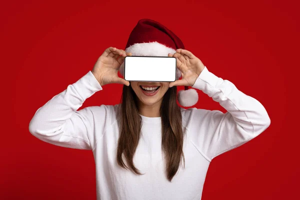 Anuncio móvil. Alegre dama usando Santa Sombrero cubriendo los ojos con Smartphone en blanco — Foto de Stock