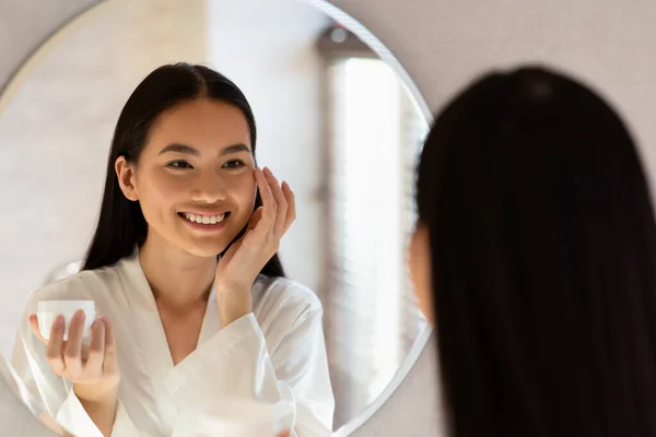 Mirror reflection of happy asian lady applying eye care product — Stock Photo, Image