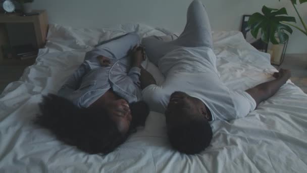 Счастливая африканская любящая пара в пижаме, отдыхающая в постели, держащаяся за руки и смеющаяся, наслаждающаяся совместным времяпрепровождением — стоковое видео