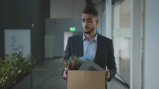 Молодого расстроенного менеджера с Ближнего Востока увольняют, он ходит по офисному зданию и несет коробку с личными вещами — стоковое видео