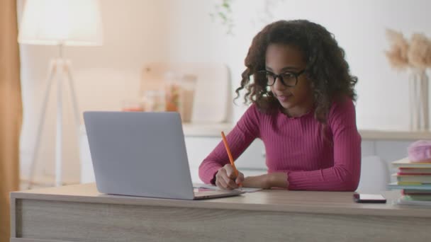 Liten afrikansk amerikansk flicka i glasögon skriva uppgift i kopiera bok, titta på främmande språk lektion på laptop på nätet — Stockvideo