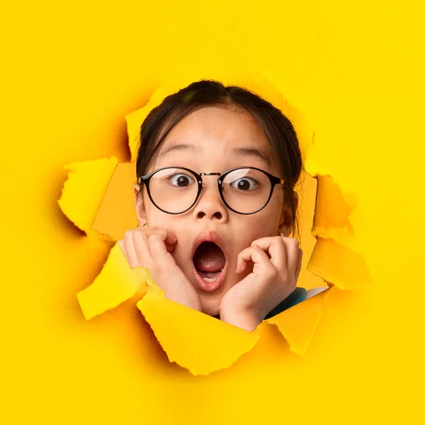 Chocado ásia menina olhando através de buraco no rasgado laranja papel, expressando excitação e surpresa — Fotografia de Stock