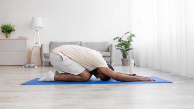 Sakallı, beyaz spor kıyafetli, paspasın altında yoga yapan Afrikalı genç bir erkek.