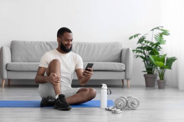 Gülümseyen bin yıllık siyah adam beyaz spor kıyafetleriyle sakallı telefonda yazan yerde oturuyor.