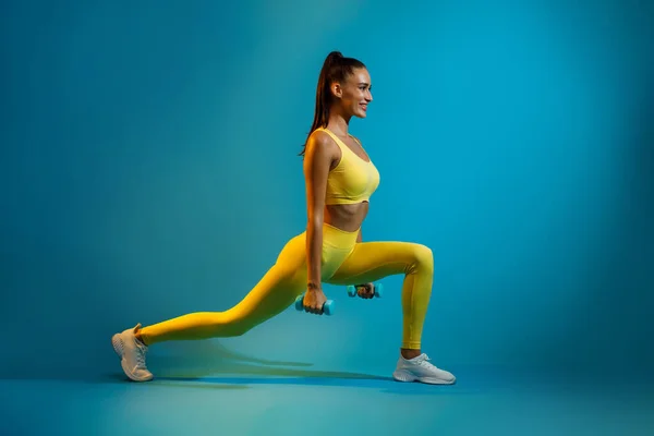 Femmina sportiva che fa esercizio profondo del affondo che tiene i manubri, sfondo blu — Foto Stock