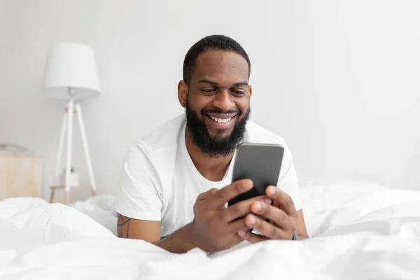 Ευτυχισμένη χιλιετή αφρικάνος Αμερικανός γενειοφόρος άνδρας βρίσκεται στο λευκό κρεβάτι στο εσωτερικό υπνοδωμάτιο και πληκτρολογώντας στο smartphone — Φωτογραφία Αρχείου