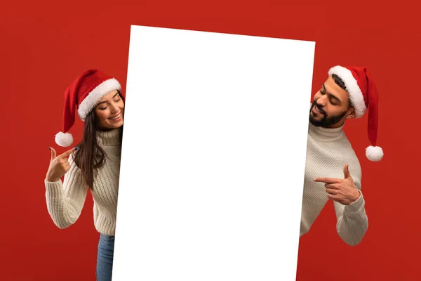 Place pour l'annonce. Heureux couple moyen-oriental dans chapeaux Santa tenant tableau blanc, regardant et pointant sur la plaque vide — Photo