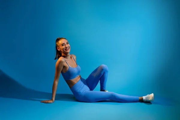 幸せなフィットネス女性が床に座っている青色の背景に微笑む — ストック写真