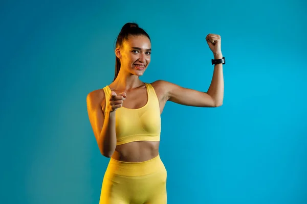 Fitness Lady mostrando bicipiti puntando dito alla macchina fotografica, sfondo blu — Foto Stock