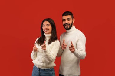 Senin sıran. Orta Doğulu mutlu çift kamerayı parmaklarıyla gösteriyor, birini seçiyor, kırmızı arka planda poz veriyor.