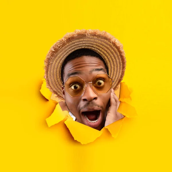 Homem negro millennial chocado gritando, usando óculos escuros e chapéu de palha, olhando através do buraco em papel laranja rasgado — Fotografia de Stock