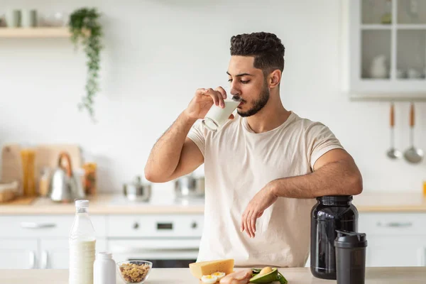 Atletische jonge Arabische man drinken eiwit shake of melk, staande in de buurt van tafel met gezonde producten in de keuken — Stockfoto