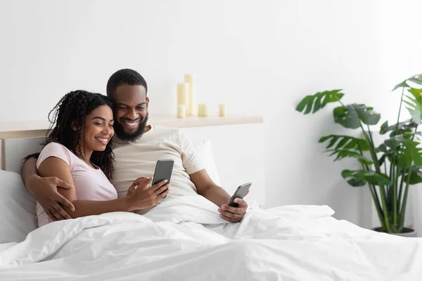 Uśmiechnięty młody afrykański mąż i żona przytulają się, patrząc na smartfony na białym łóżku — Zdjęcie stockowe