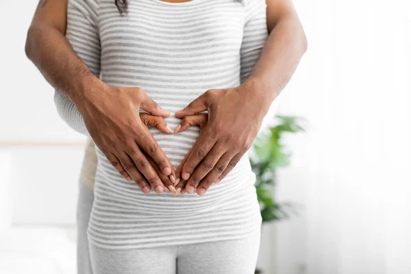 Gehakt jong Afrikaans amerikaanse man knuffels vrouw met handen in hart vorm op buik in minimalistische slaapkamer — Stockfoto