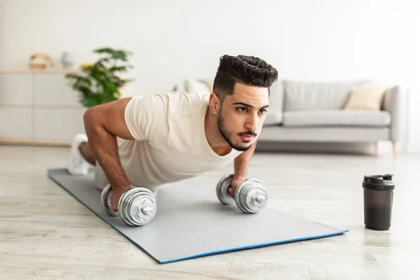 Stay home sports. Handsome young Arab man standing in plank pose with dumbbells, working out indoors — 图库照片