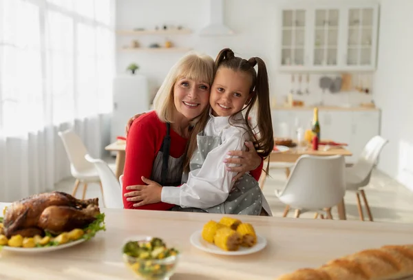 Retrato de niña alegre y su abuela en delantales cocinando la cena de Navidad, posando y abrazándose en la cocina —  Fotos de Stock