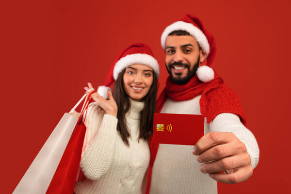 Christmas sales and shopping. Loving arab spouses holding credit card and shopper bags, wearing Santa hats — Stock Image Christmas sales and shopping. Loving arab spouses holding credit card and shopper bags, wearing Santa hats Stock Photo