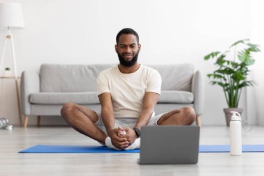 Sakin gülümseyen siyah sakallı adam yoga yapıyor, oturma odasında yerde bacak egzersizi yapıyor.