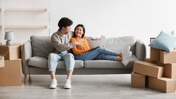 Feliz pareja asiática milenaria con portátil sentado en el sofá entre cajas de cartón y el diseño de planificación de su nuevo hogar — Foto de Stock