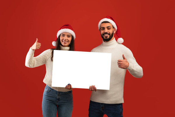 Holiday offer. Middle eastern couple in Santa hats holding blank advertisement board and showing thumbs up — Stock Image Holiday offer. Middle eastern couple in Santa hats holding blank advertisement board and showing thumbs up Stock Picture