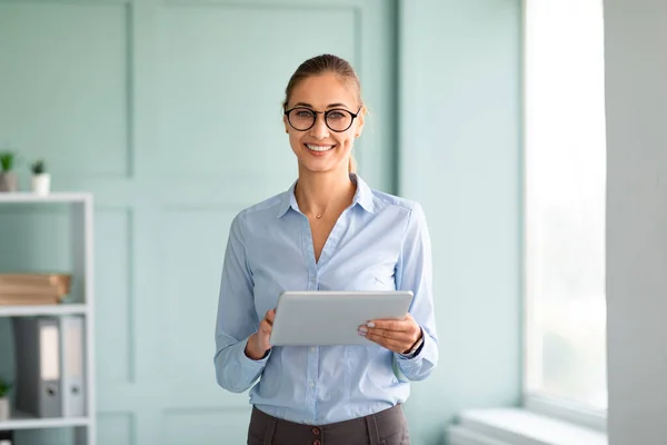 Positieve zakenvrouw in formele kleding met behulp van tablet op kantoor, kijken en glimlachen op de camera, staan in de buurt van de werkplek — Stockfoto