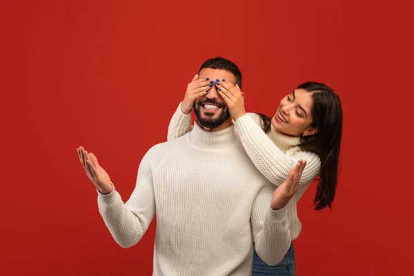 Concept de surprise romantique. Joyeux arabe femme couvrant ses yeux de petit ami de dos, fond rouge studio — Photo