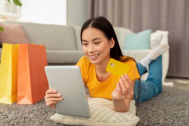 Alışveriş konsepti. Kredi kartı tutan mutlu genç Asyalı bayan, yanında hediye çantalarıyla tabletle alışveriş yapıyor.