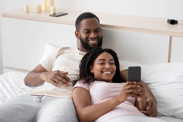 Veselý mladý africký americký manžel objetí manželka s smartphonem na bílé posteli v ložnici interiér — Stock fotografie