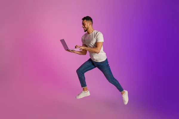 Longitud completa de alegre joven árabe utilizando ordenador portátil y saltar en luz de neón, espacio de copia — Foto de Stock