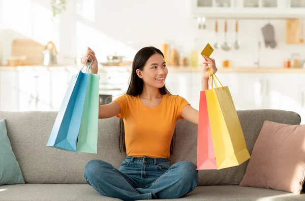 Gran venta y concepto de descuento. Feliz asiática dama sentada en sofá con coloridas bolsas de compras y tarjeta de crédito — Foto de Stock