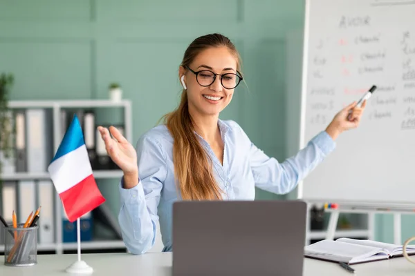 Heyecanlı bayan öğretmen internette Fransızca dersi veriyor, dizüstü bilgisayar kullanıyor, dil bilgisi kurallarını anlatıyor, iş yerinde oturuyor. — Stok fotoğraf