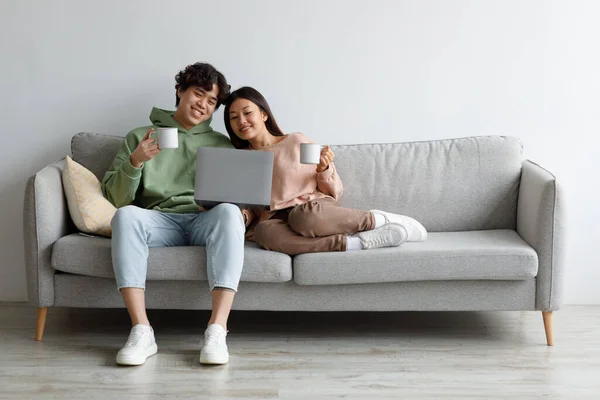 Ευτυχισμένο ασιατικό ζευγάρι κάθεται στον καναπέ με φορητό υπολογιστή και καφέ, περιήγηση στο διαδίκτυο μαζί στο σπίτι, ελεύθερο χώρο — Φωτογραφία Αρχείου