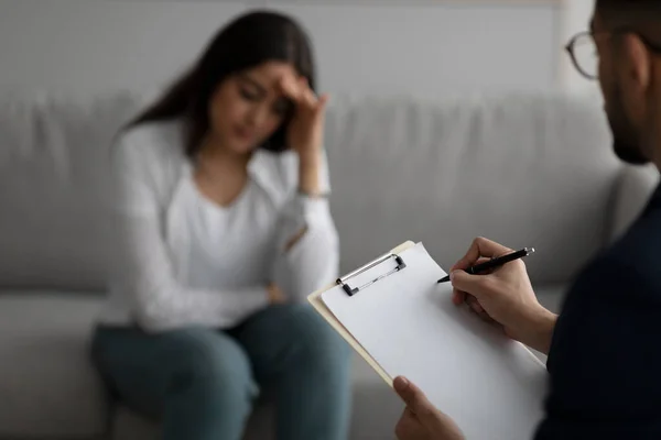 沮丧的中东女人在医务室咨询心理医生，专注于在剪贴板上写精神病医生的文章 — 图库照片
