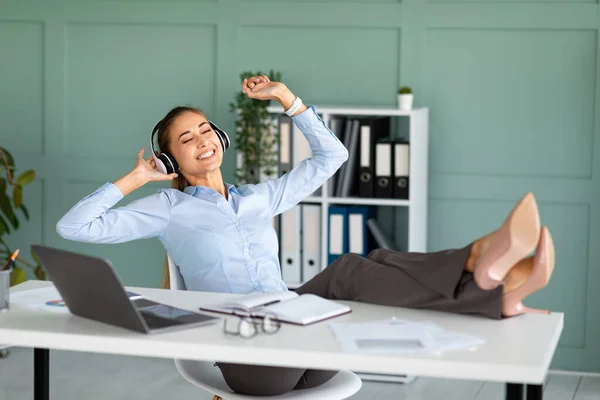 Empresária relaxada desfrutando de música em fones de ouvido, sentada em poltrona e colocando os pés na mesa — Fotografia de Stock