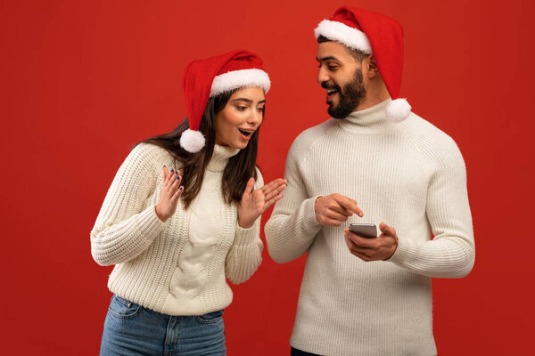 Happy arab man receiving message showing smartphone to surprised wife, wearing Santa hats, red studio background — Stock Image Happy arab man receiving message showing smartphone to surprised wife, wearing Santa hats, red studio background Royalty Free Stock Photos