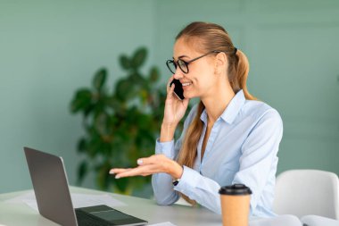 Kurumsal iletişim kavramı. İş yerinde oturup bilgisayarla uğraşırken akıllı telefondan konuşan genç bir kadın.