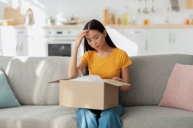 Yanlış paket. Kanepede oturan üzgün Asyalı kadın paketsiz bir kutuyla şok içinde alnına dokunuyor.