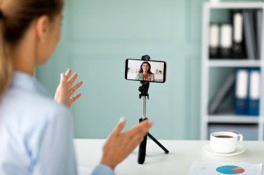 Tripodun üzerinde akıllı telefonun önünde oturan kadın öğretmen, video görüşmesi, eğitim dersi, öğrencilerle konuşma.