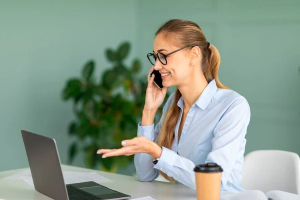 Conceito de comunicação corporativa. Jovem mulher falando no smartphone enquanto está sentado no local de trabalho e trabalhando no laptop — Fotografia de Stock