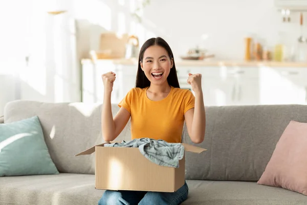 Überglückliche Asiatin packt Pappkarton-Paket aus, reagiert emotional auf erfolgreichen Einkauf, ballt die Fäuste — Stockfoto